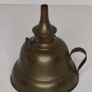 May include: A vintage brass oil lamp with a rounded base, a long spout, and a small handle. The lamp has a tarnished finish, suggesting age and use. The design is simple and functional, typical of early lighting devices.
