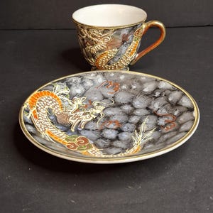 May include: A porcelain teacup and matching saucer set featuring a dark gray and white mottled background with a hand-painted orange and gold dragon motif. The cup has a gold-trimmed rim and handle, while the saucer features a gold-trimmed edge.