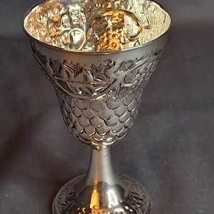 Puede incluir: Un cáliz plateado con diseños en relieve. La copa presenta una parte superior acampanada, un cuerpo texturizado y una base detallada. El interior de la copa tiene un acabado dorado, que contrasta con el exterior plateado. El cáliz se utiliza probablemente para ceremonias religiosas. Altura aproximada: 15 cm.
