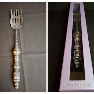 May include: A silver-toned serving fork with a four-tined head featuring a fan-shaped base and an ornate, multi-sectioned handle with decorative ridges and patterns. The fork is also shown inside a rectangular lavender box labeled STERLING SILVEE.