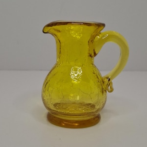 Puede incluir: Una jarra pequeña de vidrio amarillo con acabado craquelado. La jarra tiene un cuerpo redondeado, un asa curva y un pico para verter. La base es de un tono amarillo más oscuro. Esta jarra decorativa es adecuada para servir líquidos.