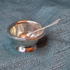 May include: A small, silver-colored salt cellar with a matching spoon. The bowl of the cellar is round and sits on a small pedestal base. The spoon rests inside the bowl, ready for use. The items are reflective and appear to be made of metal.