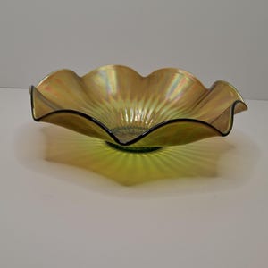 May include: An iridescent, ruffled-edge glass bowl with a golden hue. The bowl has a scalloped rim and a dark rim. The bowl's base is a dark green color. The bowl is likely used for decorative purposes.