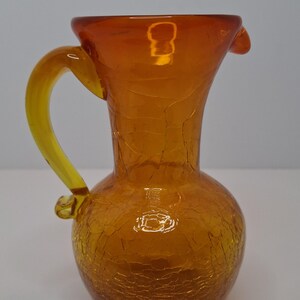 May include: An amber glass pitcher with a crackle effect. The pitcher has a rounded base, a flared top, and a yellow handle. The spout is slightly curved. The pitcher is likely vintage and suitable for decorative use.