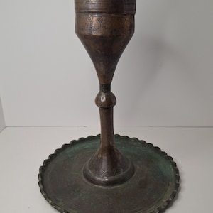 May include: An antique bronze candle holder with a wide, scalloped base and a tall, conical body. The body has decorative etched patterns near the top. The metal has a dark, aged patina with hints of green oxidation, suggesting its age. The candle holder is designed to hold a single candle.