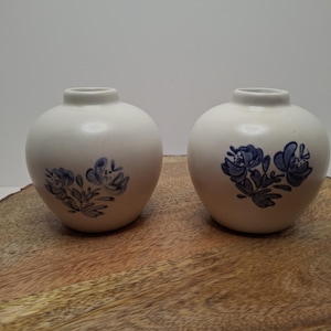 May include: Two small, round, off-white ceramic vases. Each vase features a blue floral design. The vases are displayed on a wooden surface against a plain white background. The vases have a small opening at the top.