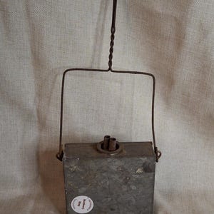 May include: A rectangular, galvanized metal container with a weathered, mottled gray finish. It features a top-mounted wire handle with a twisted design and a central opening with two small, protruding metal tubes. A circular white sticker reads The Collection of Matthew J. King.