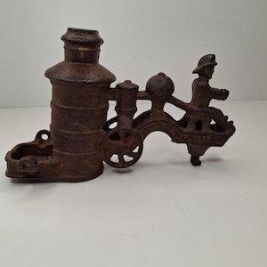 May include: A vintage cast iron match holder and ashtray in the shape of a train. The brown piece features a cylindrical body, a small tray, a wheel, and a figure of a person. The piece has a rustic, aged appearance.