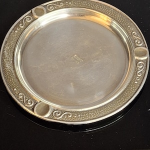 Pode incluir: Um prato redondo prateado com uma borda decorativa. A borda apresenta um padrão texturizado e desenhos de volutas estilizadas. O centro do prato é liso e refletor. O prato parece ser de metal.
