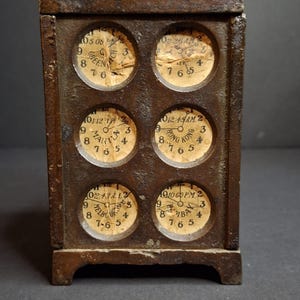 May include: A rectangular, weathered metal world clock featuring six circular analog clock faces arranged in two columns. Each face displays a different city name, including Greenwich, Manila, Hong Kong, Melbourne, and Bombay, with aged paper dials.