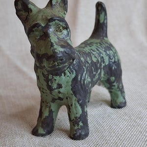 May include: A small, weathered figurine of a dog, primarily in shades of green and black. The dog is standing with its tail up and ears perked. The paint has a distressed look, adding to its vintage charm. The figurine is likely made of ceramic or a similar material.
