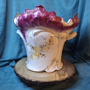 May include: A ceramic planter featuring a white base with yellow floral patterns and gold trim accents. The top rim is shaped with a deep magenta, ruffled edge. The vessel includes small side handles and a curved, ornate body design.