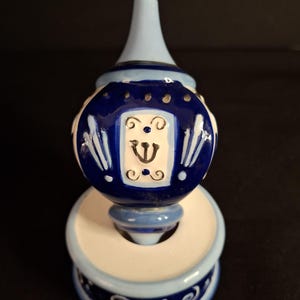 May include: A ceramic bell-shaped ornament with a pointed top, featuring a white base and deep blue accents. The center displays a rectangular panel with a black trident-like symbol, surrounded by white scrollwork and blue painted details.