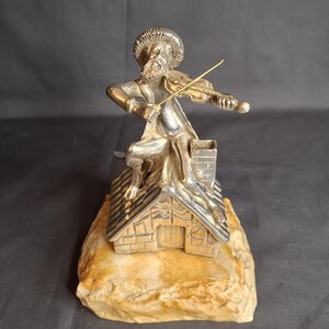 May include: A silver-toned figurine of a bearded person wearing a hat, sitting on a roof and playing a violin with a gold-toned bow. The structure features a chimney and is mounted on a textured, light brown, rectangular stone-like base.