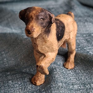 Op de afbeelding: Een kleine, vintage hondenfiguur met een zwarte en bruine kop en een zwarte vlek op zijn rug. De hond is bruin en lijkt gemaakt te zijn van een composietmateriaal. Het beeldje staat op een getextureerd, blauwgroen oppervlak.
