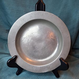 May include: A round, silver-colored plate with a slightly raised rim, resting on a black display stand. The plate has a shallow center and appears to be made of metal. The stand has a curved design.