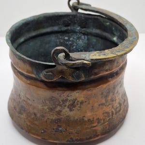 May include: A small, antique copper bucket with a curved handle. The bucket has a rounded body with a slightly flared rim and a handle attached with a decorative loop. The copper has a weathered, aged appearance with areas of green oxidation.