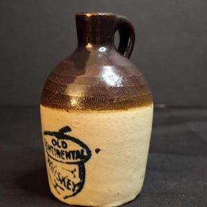 May include: A small stoneware jug with a brown glazed neck and handle, featuring a cream-colored body. The front displays a hand-drawn blue ink illustration of an acorn containing the text OLD CONTINENTAL WHISKEY in a bold, dark blue font.