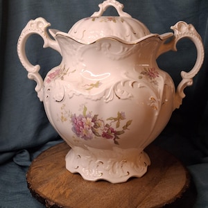 May include: A white porcelain lidded tureen featuring two ornate handles, a scalloped rim, and a base with embossed scrollwork. The body and lid are decorated with a delicate floral pattern in shades of pink, purple, and green, accented with gold trim.