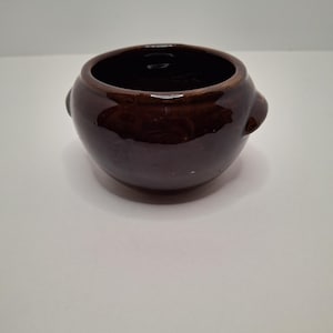 Könnte beinhalten: Eine kleine, runde, braune Keramikschale mit zwei kleinen Griffen an den Seiten. Die Schale hat eine glänzende Oberfläche und ein dunkles Inneres. Die Schale dient wahrscheinlich zum Servieren oder Aufbewahren.