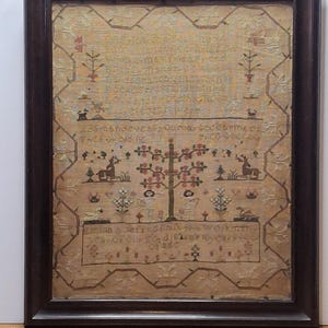 以下が含まれることがあります： 濃い茶色の木製フレームに入ったアンティークのサンプラー。サンプラーには、木、花、動物などの刺繍されたテキストと装飾要素が施されています。テキストには「Emma Jeffs」という名前と「1824」という日付が含まれています。