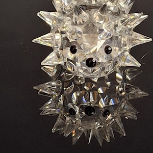 Puede incluir: Una figura de erizo de cristal transparente y facetado con ojos de cuentas negras y una nariz negra. El cuerpo del erizo está cubierto de púas de cristal puntiagudas. La figura se refleja en una superficie oscura.