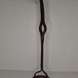 May include: A tall, dark brown wrought iron candle holder. It has a circular base, a central stem with a diamond-shaped detail, and a curved top for a candle. The textured finish and design suggest an antique or rustic aesthetic. The height is approximately 100 cm.
