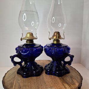 Puede incluir: Dos lámparas de aceite azul cobalto antiguas con chimeneas de vidrio transparente. Las lámparas tienen bases ornamentadas con asas y accesorios de latón. Las lámparas se exhiben sobre una base rústica redonda de madera.