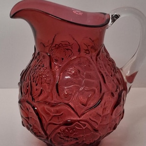 May include: A cranberry-colored glass pitcher with a clear handle. The pitcher features a detailed embossed floral design. The handle is clear glass. The pitcher has a spout for easy pouring.