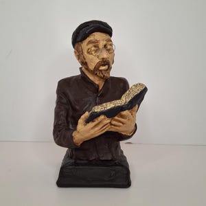 Könnte beinhalten: Büste eines Mannes mit Bart, Brille und Mütze, der ein offenes Buch hält. Die Figur trägt eine dunkelbraune Jacke. Der Sockel hat eine dunkle, rechteckige Form. Das Buch hat goldfarbene Seiten.
