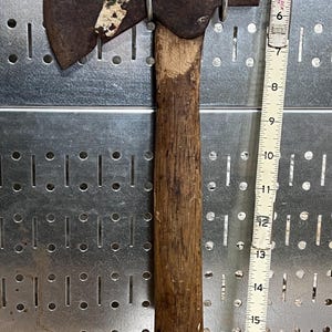 May include: A vintage hatchet with a wooden handle and a rusty metal head. The hatchet is approximately 16 inches long. The handle shows signs of wear, and the metal head has a worn appearance. The hatchet is displayed against a metal pegboard.