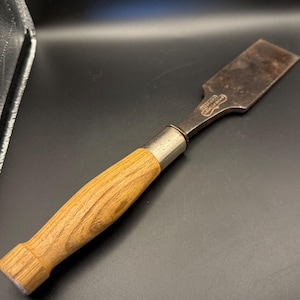 May include: A woodworking chisel with a wooden handle and a metal blade. The handle is light brown, and the blade is a darker, aged metal. The chisel has a metal ferrule where the handle meets the blade. The tool is designed for shaping wood.