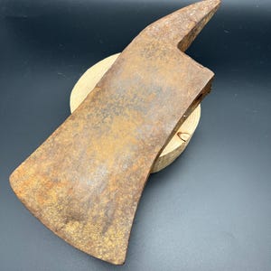 May include: A weathered, rusted metal axe head with a wide, curved cutting edge and a pointed poll. The heavy iron tool displays significant oxidation and surface texture, showing signs of age and past use, resting on a small, circular wooden base.