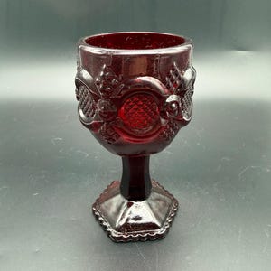 May include: A ruby red glass goblet with ornate embossed floral and geometric patterns. The goblet features a hexagonal base and a detailed stem. The bowl of the glass has a textured design, adding to its vintage appeal.