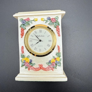 May include: Cream-colored quartz clock with a gold-tone bezel and Roman numeral hour markers. The clock face reads "Reflections." The clock is decorated with colorful floral arrangements and red ribbon accents.