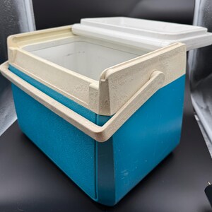 May include: A vintage blue and off-white cooler with a white lid. The cooler has a handle and is made of a durable plastic material. The lid is partially open, revealing the interior. The cooler is designed to keep items cold.