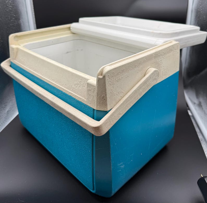 May include: A vintage blue and off-white cooler with a white lid. The cooler has a handle and is made of a durable plastic material. The lid is partially open, revealing the interior. The cooler is designed to keep items cold.