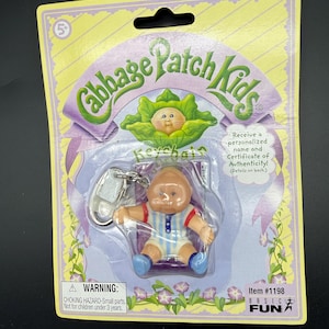 May include: Cabbage Patch Kids keychain in original packaging. The doll has a tan complexion, blue eyes, and is wearing a blue and white striped outfit. The packaging is yellow and purple with the Cabbage Patch Kids logo.