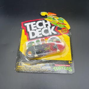 May include: A Tech Deck fingerboard featuring a Santa Cruz and Teenage Mutant Ninja Turtles collaboration graphic. The miniature skateboard has a black and red deck with purple and green character artwork, encased in original yellow and black packaging.