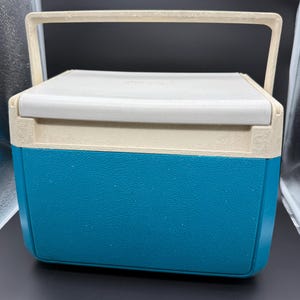 May include: A vintage Coleman cooler with a white lid and handle, a beige trim, and a teal blue base. The cooler has a rectangular shape and a handle for easy carrying. The cooler is designed to keep items cold.