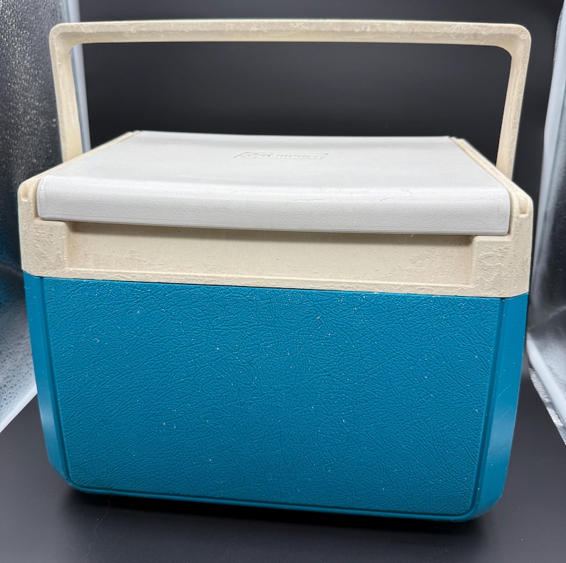 May include: A vintage Coleman cooler with a white lid and handle, a beige trim, and a teal blue base. The cooler has a rectangular shape and a handle for easy carrying. The cooler is designed to keep items cold.
