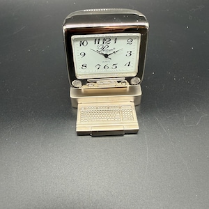 May include: A silver-toned quartz clock shaped like a computer monitor, with a keyboard base. The clock face has black numerals and hands, with the brand name "Platinum" visible. The clock is approximately 4 inches tall.