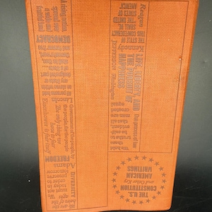May include: A rectangular orange book cover featuring embossed text and a circular star pattern. The text includes names like Reagan, Kennedy, Jefferson, Washington, Lincoln, Eisenhower, and Adams, alongside phrases such as The U.S. Constitution and Life, Liberty, and the Pursuit of Happiness.