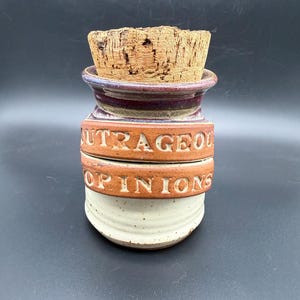 May include: A ceramic jar with a cork stopper featuring a two-tone glaze. The upper section is a deep purple, while the lower section is a speckled cream. Two raised terracotta-colored bands wrap around the center with the text OUTRAGEOUS OPINIONS.