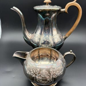 May include: A silver tea set with ornate floral detailing. The set includes a teapot with a wooden handle and a creamer. The teapot has a curved spout and a lid with a small wooden knob. The creamer has a small handle and spout.
