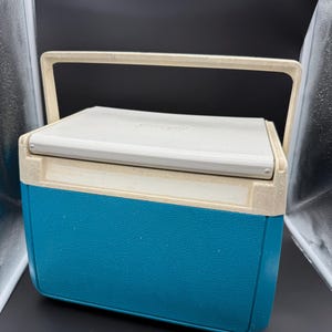 May include: A vintage teal and off-white cooler with a cream-colored handle. The cooler has a rectangular shape with a textured surface. The lid is off-white and has a slight curve. The handle is arched and made of the same cream-colored material.