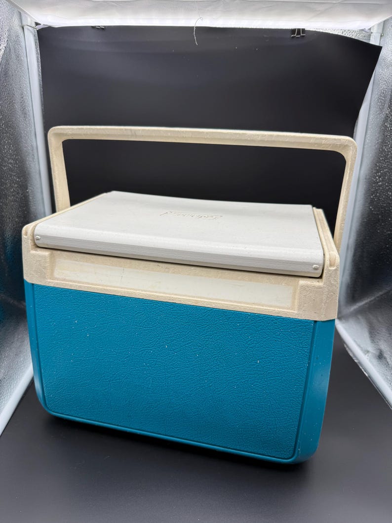 May include: A vintage teal and off-white cooler with a cream-colored handle. The cooler has a rectangular shape with a textured surface. The lid is off-white and has a slight curve. The handle is arched and made of the same cream-colored material.