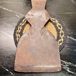 May include: A vintage, rusty metal axe head with a cross-shaped design. The axe head is resting on a round, woven coaster with black, tan, and gold colors. The axe head has a worn, aged appearance.