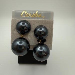 May include: A pair of vintage dark gray Carotee earrings. Each earring features two large, glossy, spherical beads suspended from a small gold-toned connector. The earrings are displayed on a beige card with the brand name "Carotee" in gold lettering.