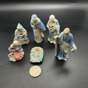 May include: A porcelain nativity set featuring six small figurines including a central infant in a manger, a kneeling figure, and four standing figures in blue, tan, and pink robes with gold accents, shown next to a US quarter for scale.
